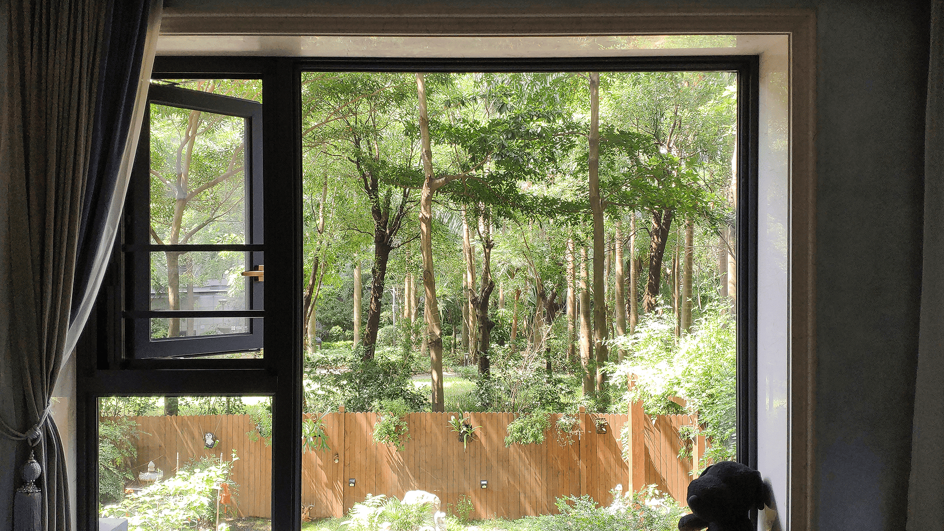 Interior view of a LIKI black aluminum casement window pushed open for ventilation, featuring an integrated insect screen and framing a garden view.