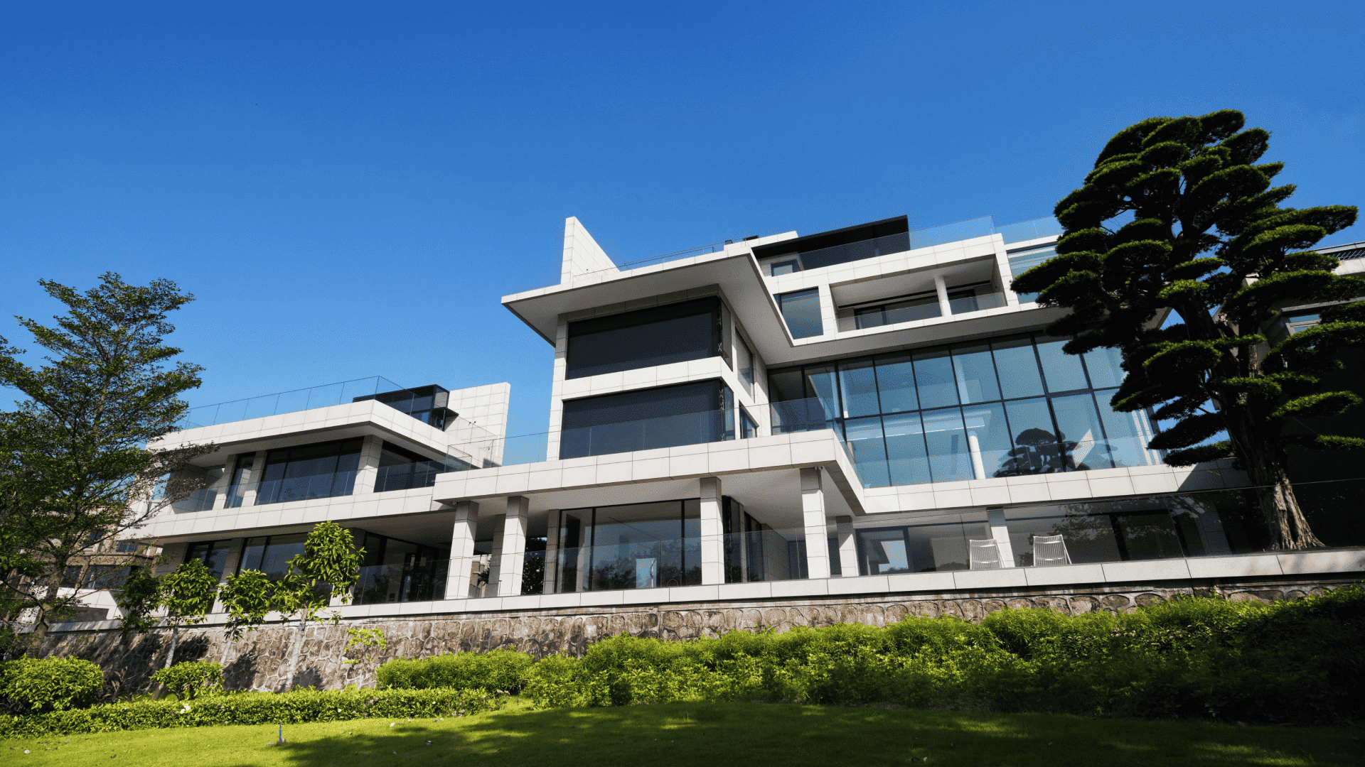 Exterior view of a contemporary lakefront villa under a clear blue sky, highlighting the massive custom Liki glass facades, clean white stone cladding, and lush landscaping that seamlessly blends the minimalist architecture with nature.